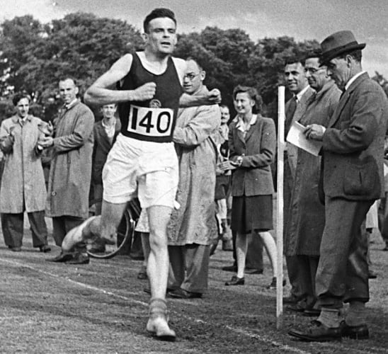 Imagem de Alan Turing, um homem caucasiano e de cabelos escuros, com trajes de corredor e o número 140 na camiseta preta, correndo e várias pessoas ao redor observando sua chegada.