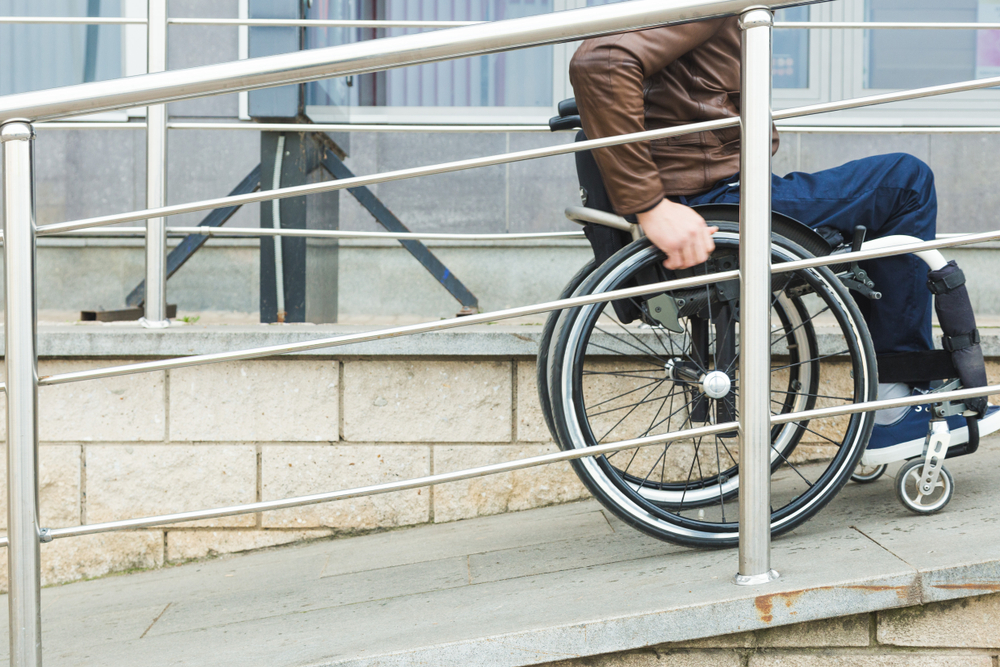 Homem de jaqueta marrom e calças azuis subindo, com uma cadeira de rodas, uma rampa cercada por corrimões metálicos e prateados.