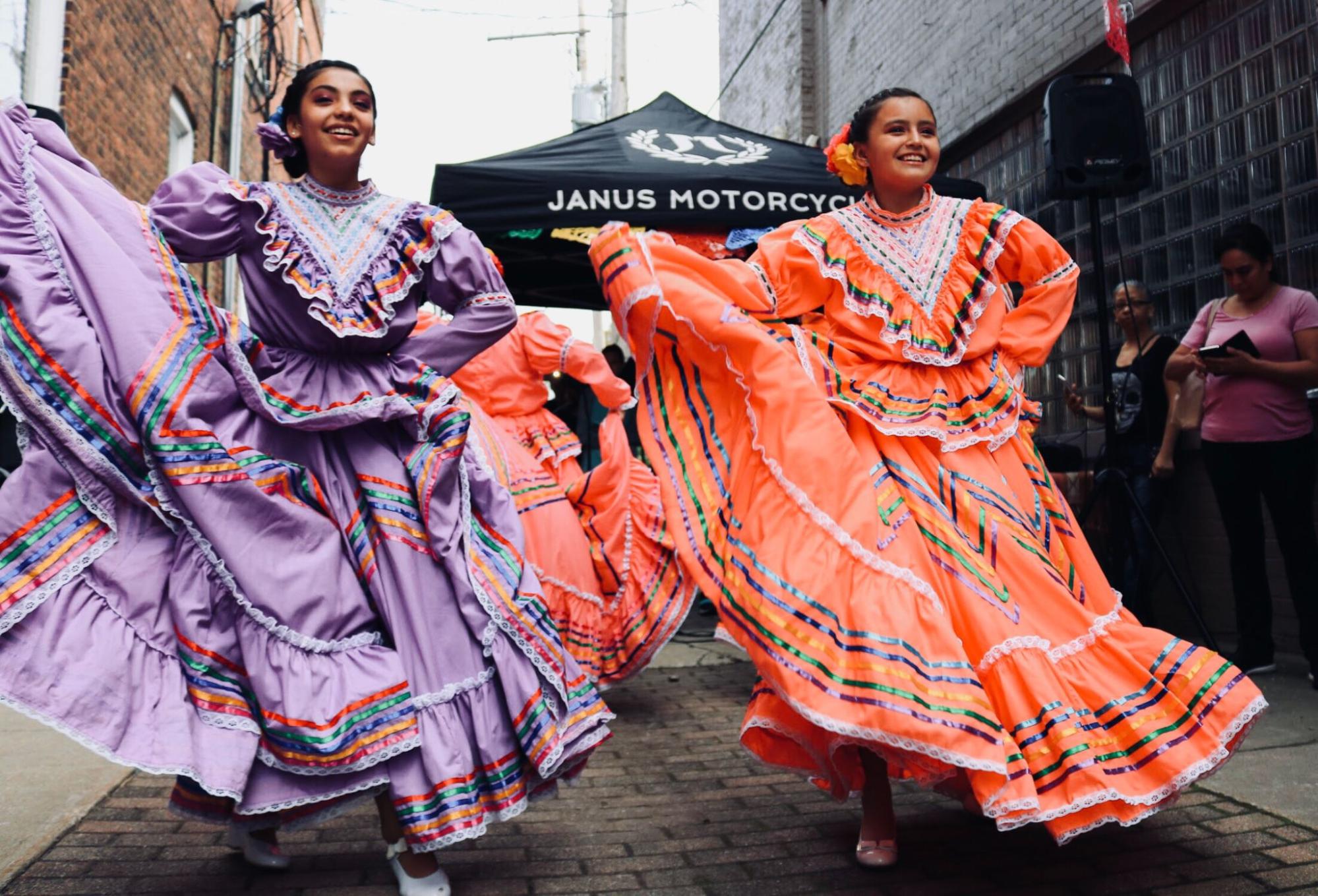 alt text: Foto de duas mulheres com vestidos longos, um roxo e outro laranja, e dançando uma dança típica de sua cultura