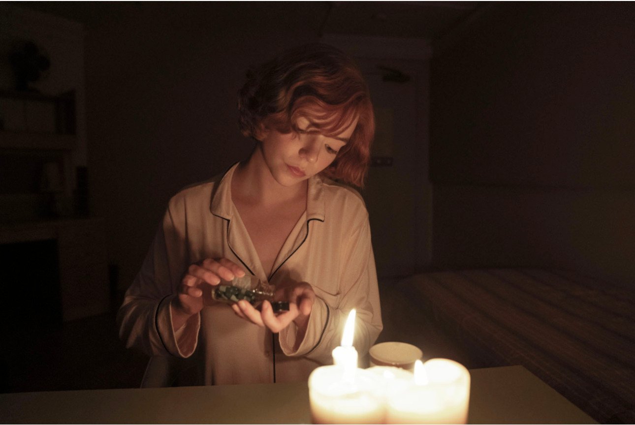 Mulher segurando um frasco de remédios em um quarto escuro em frente a uma mesa sendo iluminada por velas.