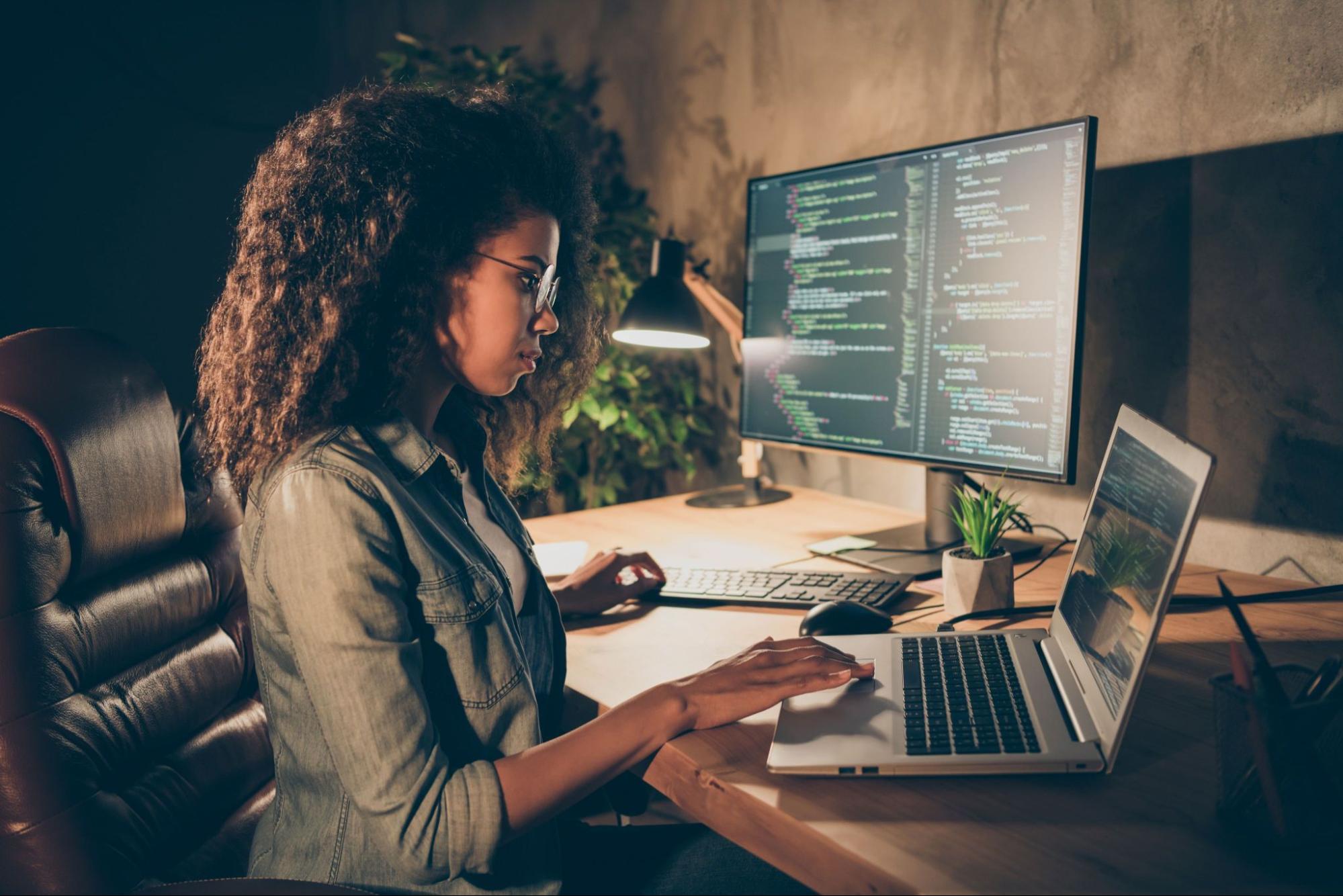 Imagem de uma mulher como pessoa desenvolvedora. À sua frente, encontra-se um notebook. Ao lado, um monitor exibindo uma IDE com um código de projeto em tela.