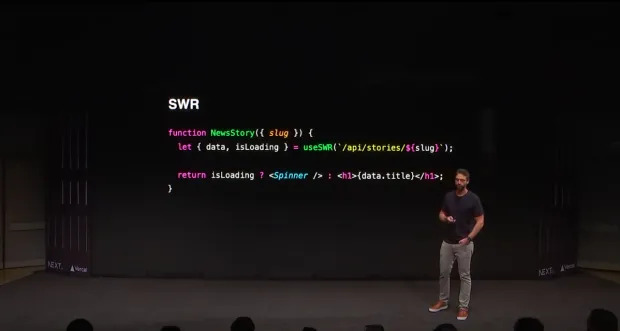 Sam Selikof está de pé em um palco ao lado de uma projeção de uma função de código de programação chamada 'NewsStory'. O código mostra o uso do gancho 'useSWR' para buscar dados de uma API com uma variável 'slug'. Ele verifica se o conteúdo está carregando e, caso esteja, mostra um componente 'Spinner'; se não, exibe o título da história em um elemento de cabeçalho 'h1'. O fundo é escuro e o logo da NEXT é visível no canto inferior esquerdo.
