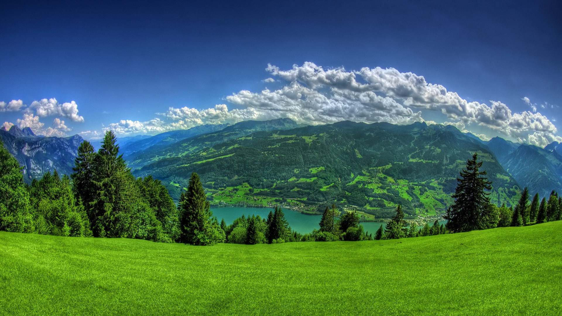 Imagem que mostra uma paisagem. No primeiro plano, há um gramado, no segundo, árvores e um lago e ao fundo, montanhas e nuvens no céu.