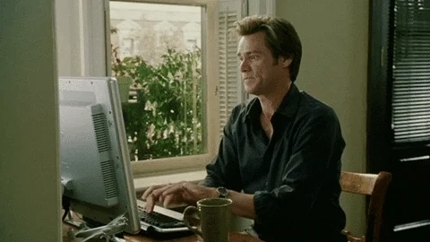 Personagem do ator Jim Carrey digitando furiosamente em um computador de mesa. O ator usa uma camisa social escura, está em uma cadeira de madeira em um local próximo a uma janela. O computador é branco e em cima da mesa está uma caneca na cor verde musgo.