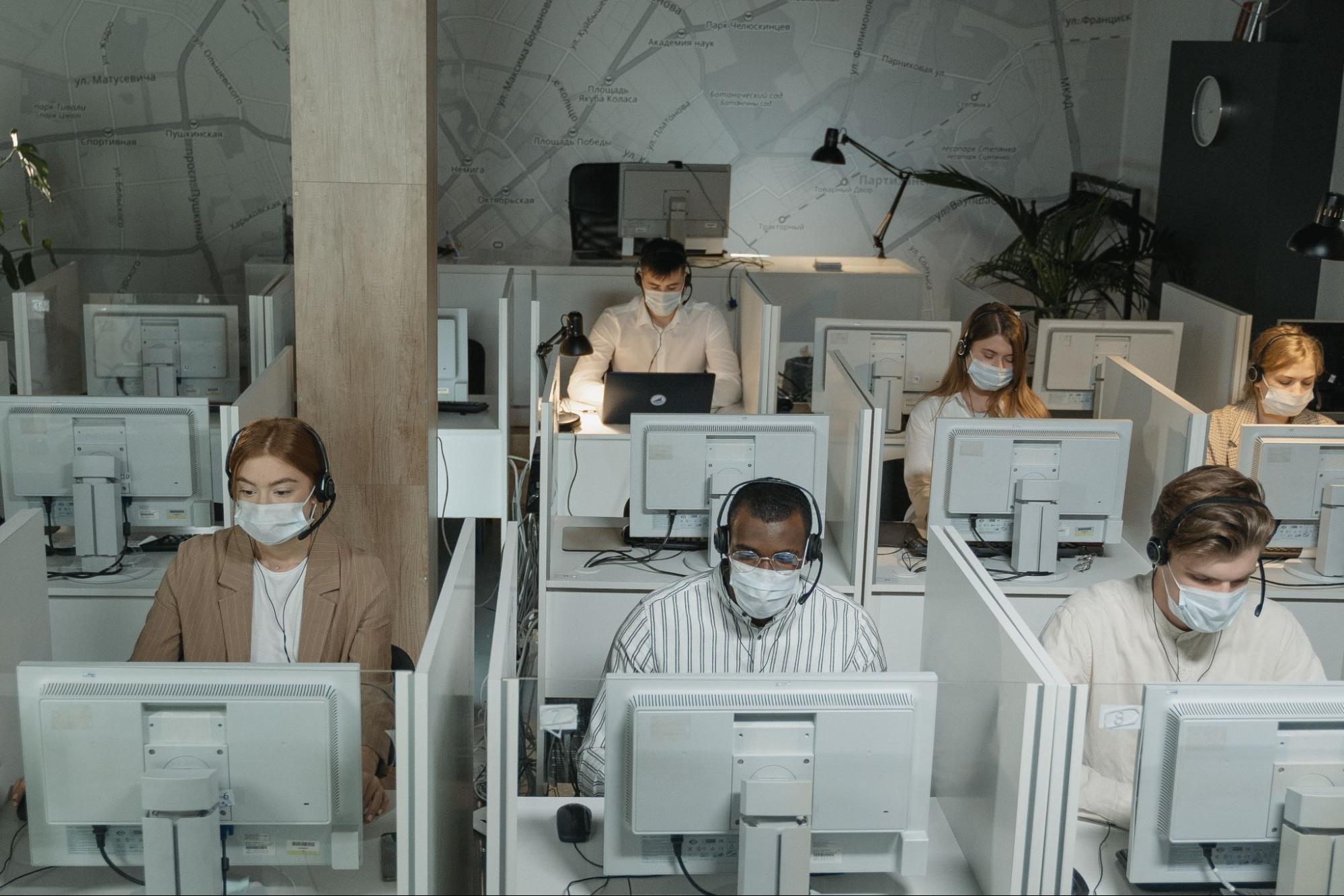 Seis pessoas trabalhando em “cabines” separadas, cada uma com o seu computador, fone de ouvido e máscara