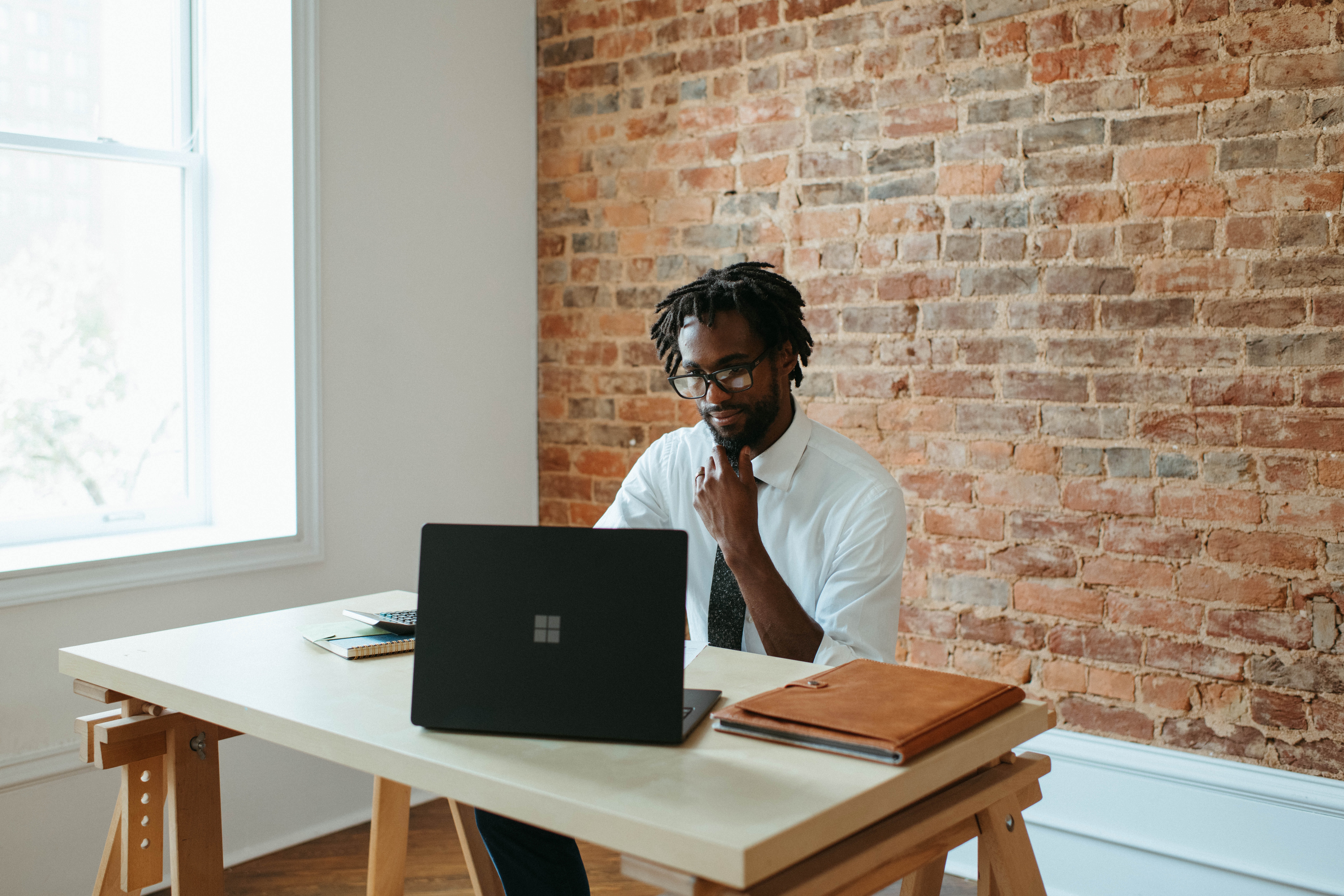 Imagem de um homem sentado de frente para uma mesa utilizando um notebook, com uma pasta, uma calculadora e um caderno ao redor do aparelho.