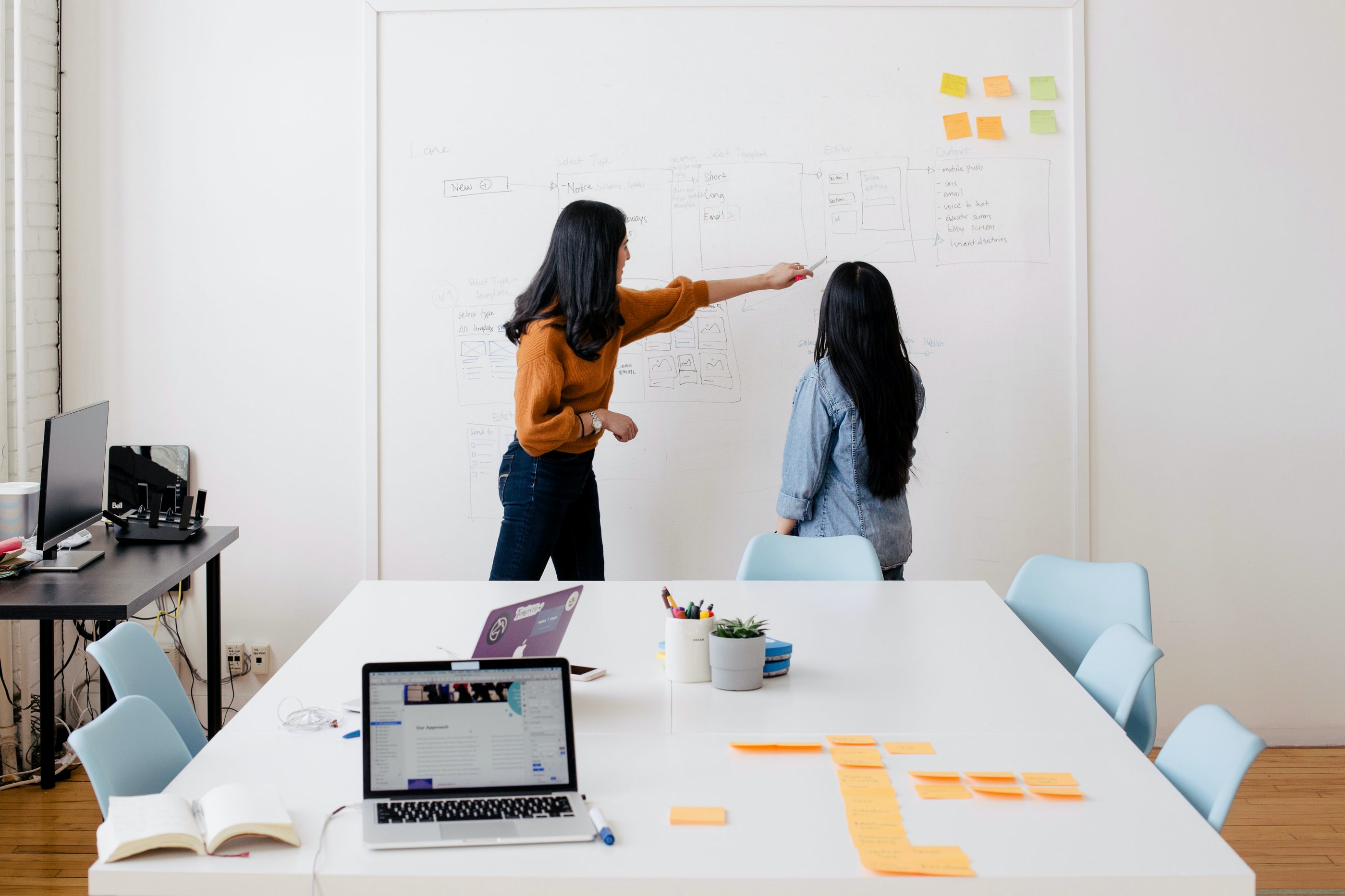 Imagem de duas pessoas em uma sala de reuniões fazendo anotações em um quadro branco. A sala é composta por duas mesas, uma pequena e outra grande, seis cadeiras, três laptops e alguns lápis, post-its e livros espalhados.