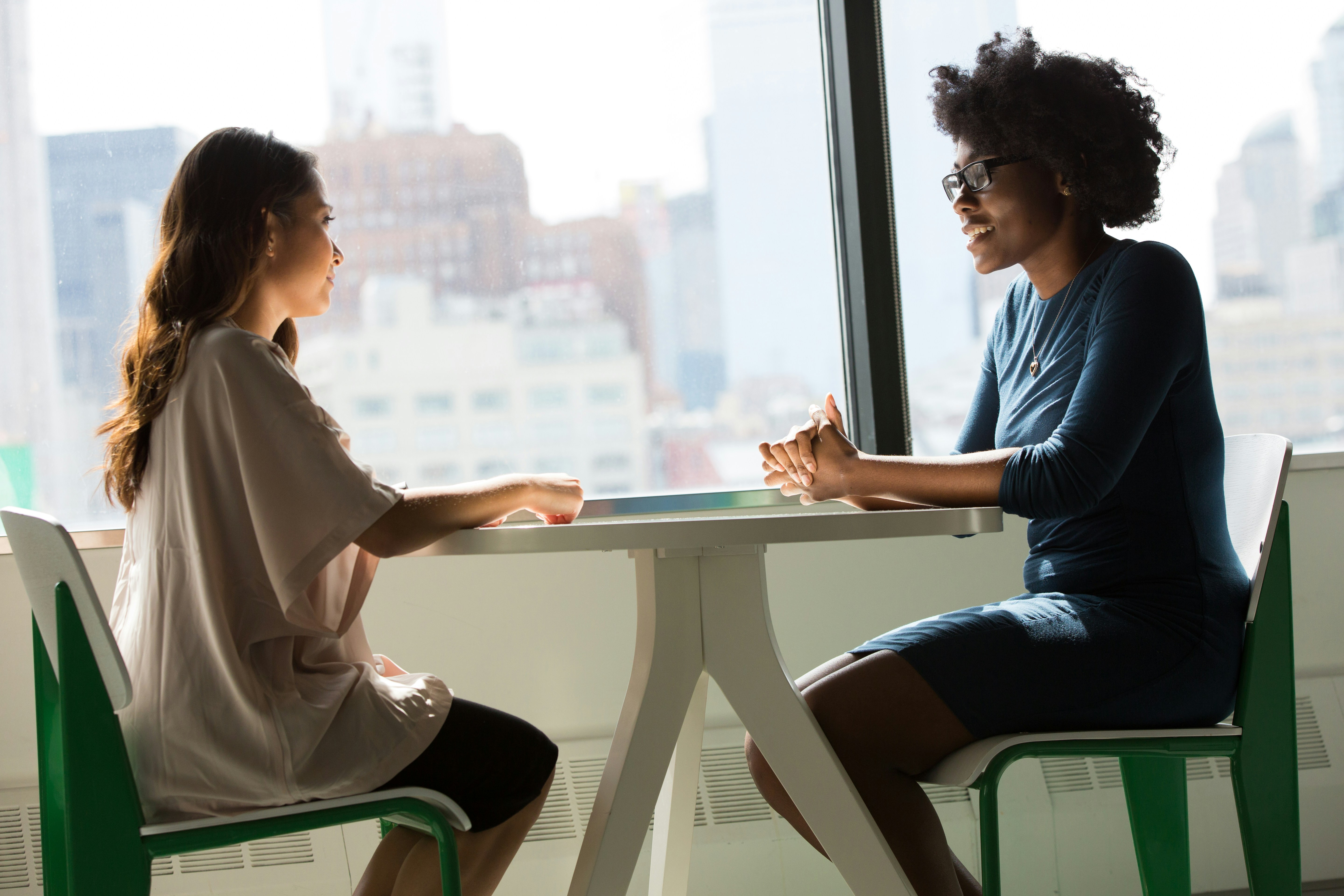 Na imagem é possível ver duas mulheres jovens sentadas em uma mesa, uma de frente para outra, representando uma conversa