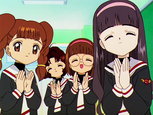 Quatro garotas em uniformes escolares, em estilo anime, batendo palmas com expressões felizes e satisfeitas. A cena transmite um sentimento de aprovação e entusiasmo.