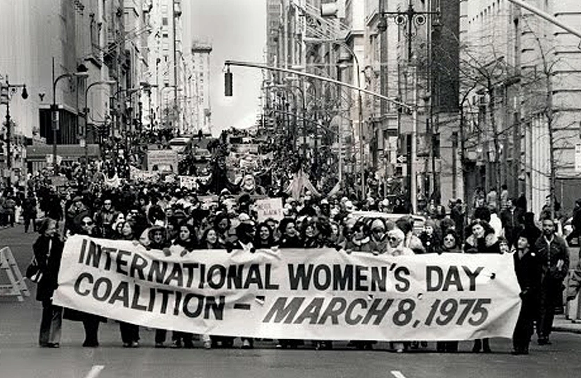 Imagem de uma manifestação de mulheres que seguram uma faixa escrito em inglês “Aliança Dia Internacional das Mulheres - 8 de março de 1975”