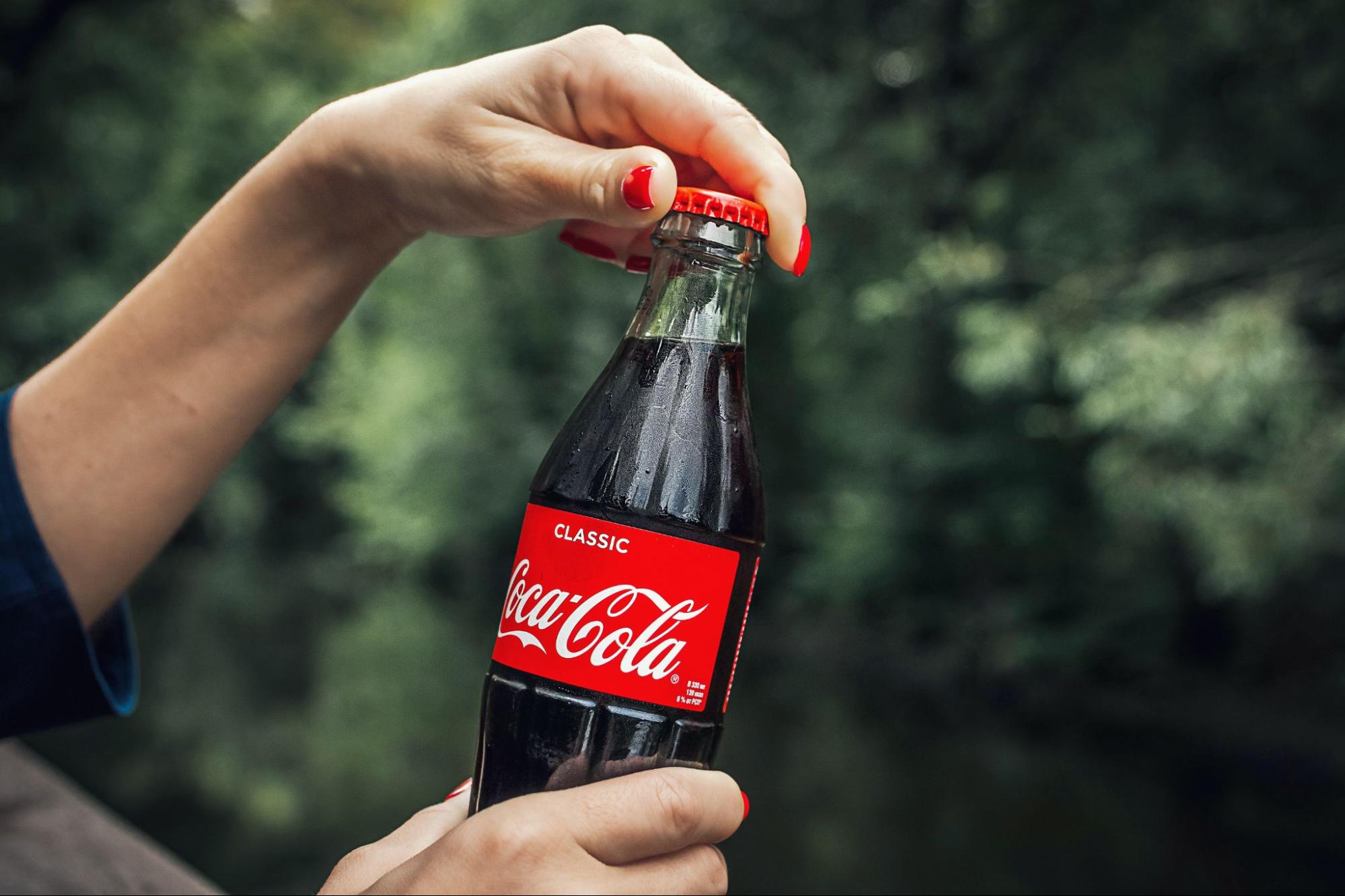 Uma foto mostrando as mãos de uma pessoa abrindo uma garrafa de Coca-Cola.