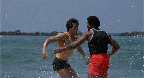 Cena do filme “Rocky”. O vídeo mostra o personagem Rocky, caucasiano e o personagem Apollo, negro. Ambos comemoraram e se abraçam numa praia, estão com shorts e blusas curtas, típicas para a prática de exercícios.
