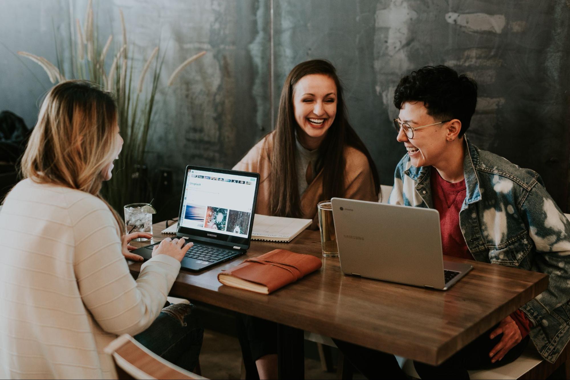 Três pessoas conversando sentadas à mesa, duas utilizando notebooks e uma utilizando um caderno.