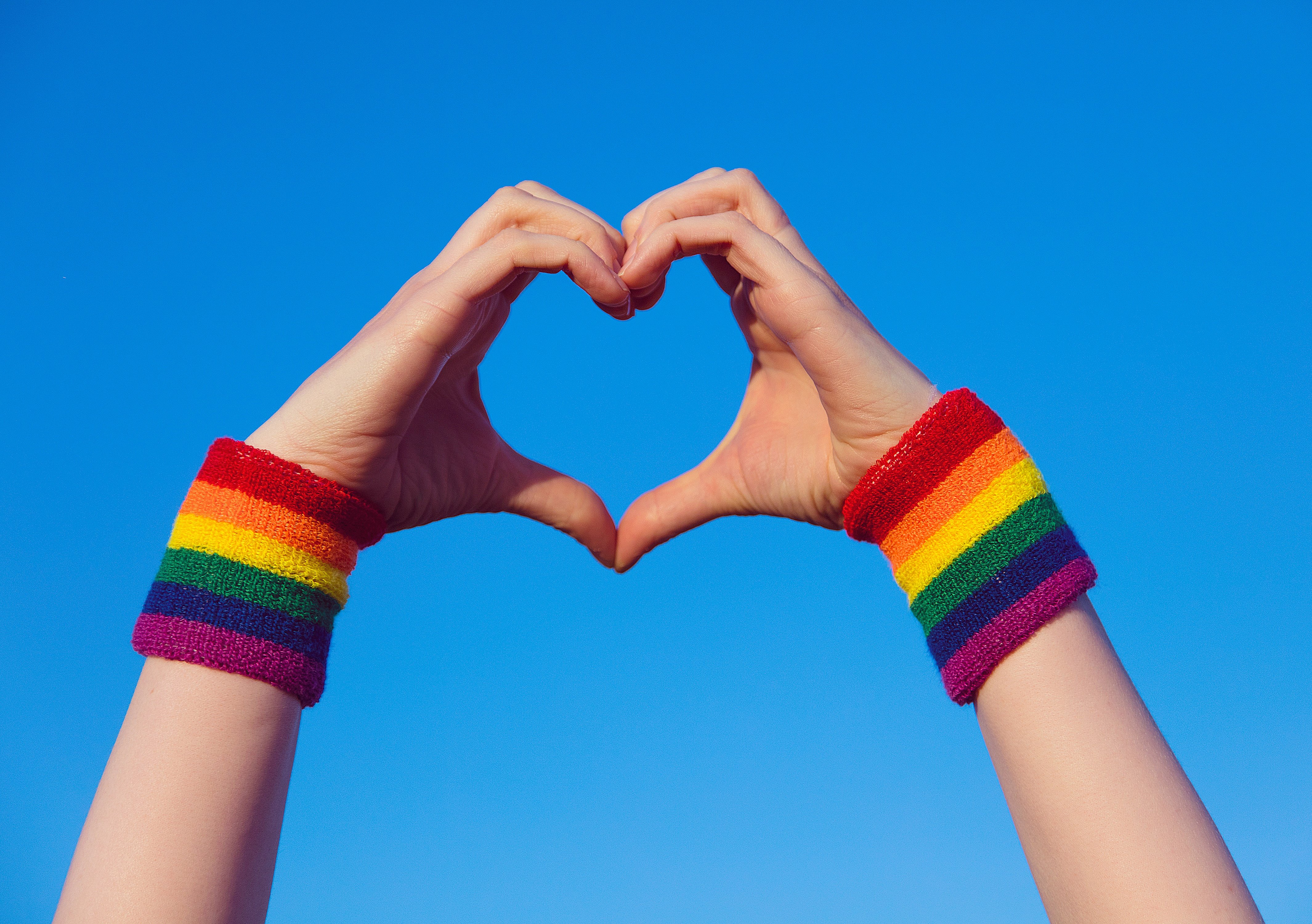 Uma pessoa fazendo coração com as mãos e nos pulsos a bandeira LGBTQIA+.