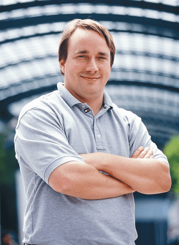 Foto do Linus Torvalds, ele está vestindo uma camisa cinza e tem os braços cruzados, exibindo um sorriso.