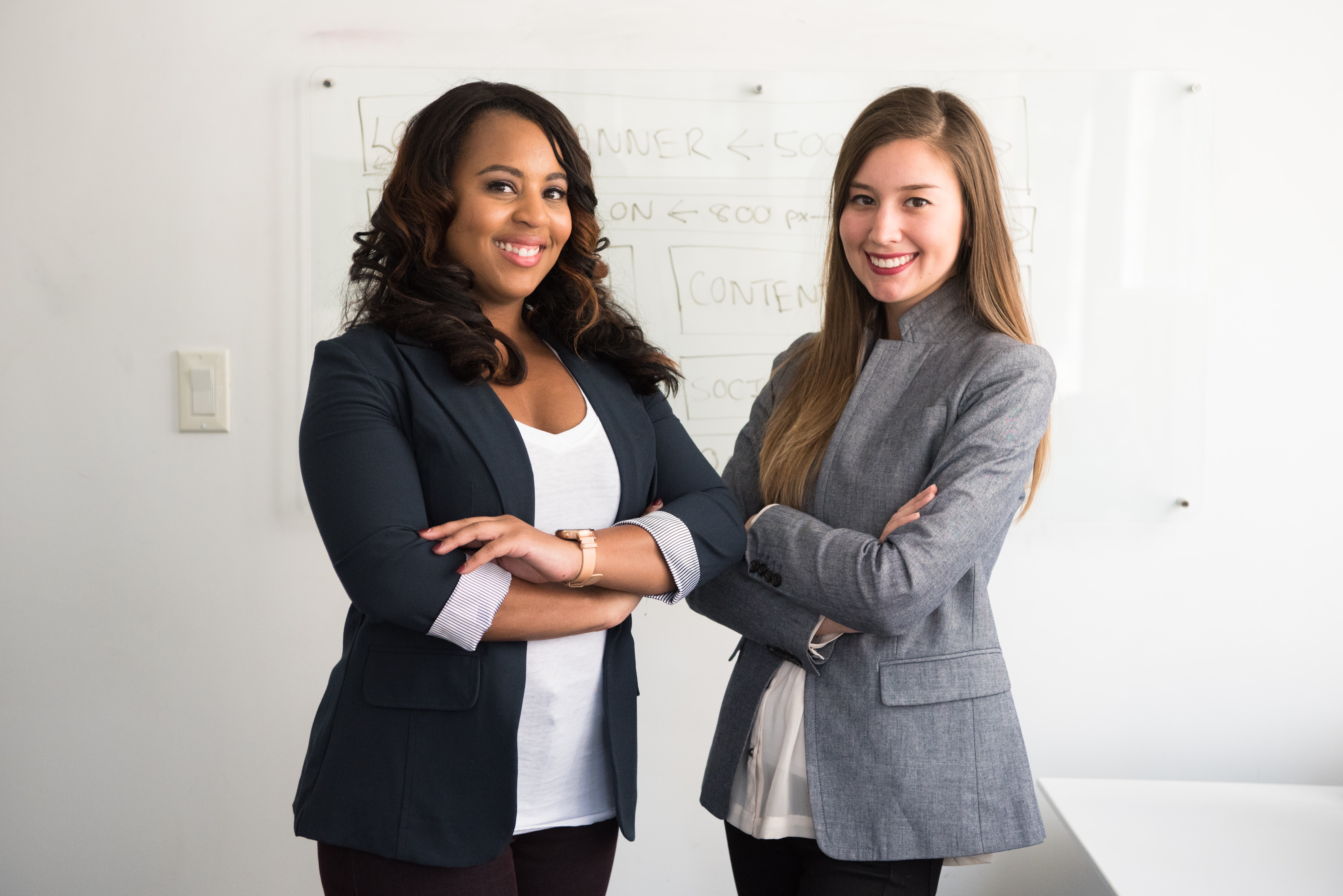 Imagem de duas mulheres em um escritório e sorrindo para a pessoa que está tirando a foto.