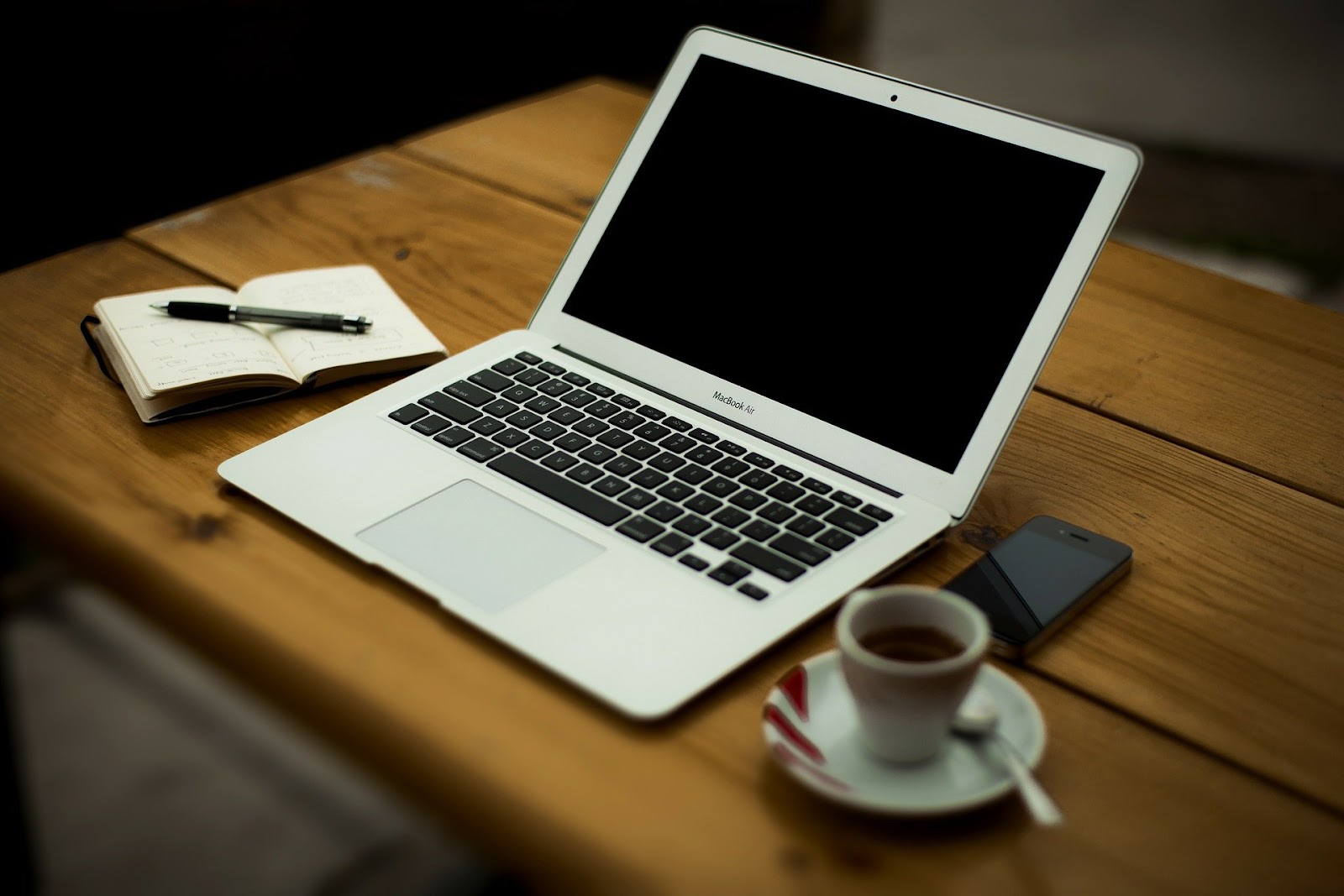 Um caderno com uma caneta em cima, uma xícara com café, um celular e um computador em cima de uma mesa de madeira.