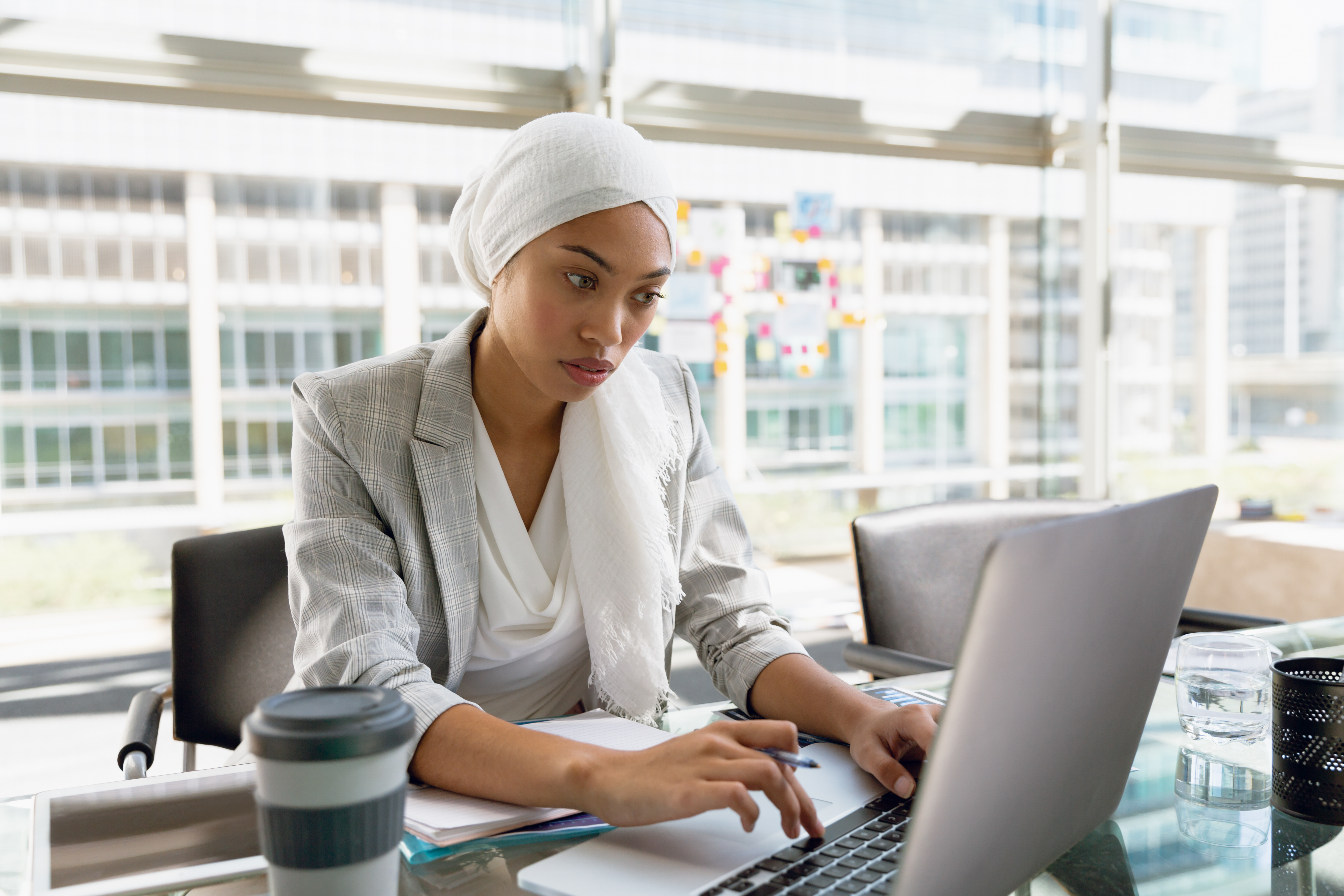 Mulher com hijab na cabeça, usando notebook à sua frente, tendo na mesa copo de café, porta lápis e copo d’água.