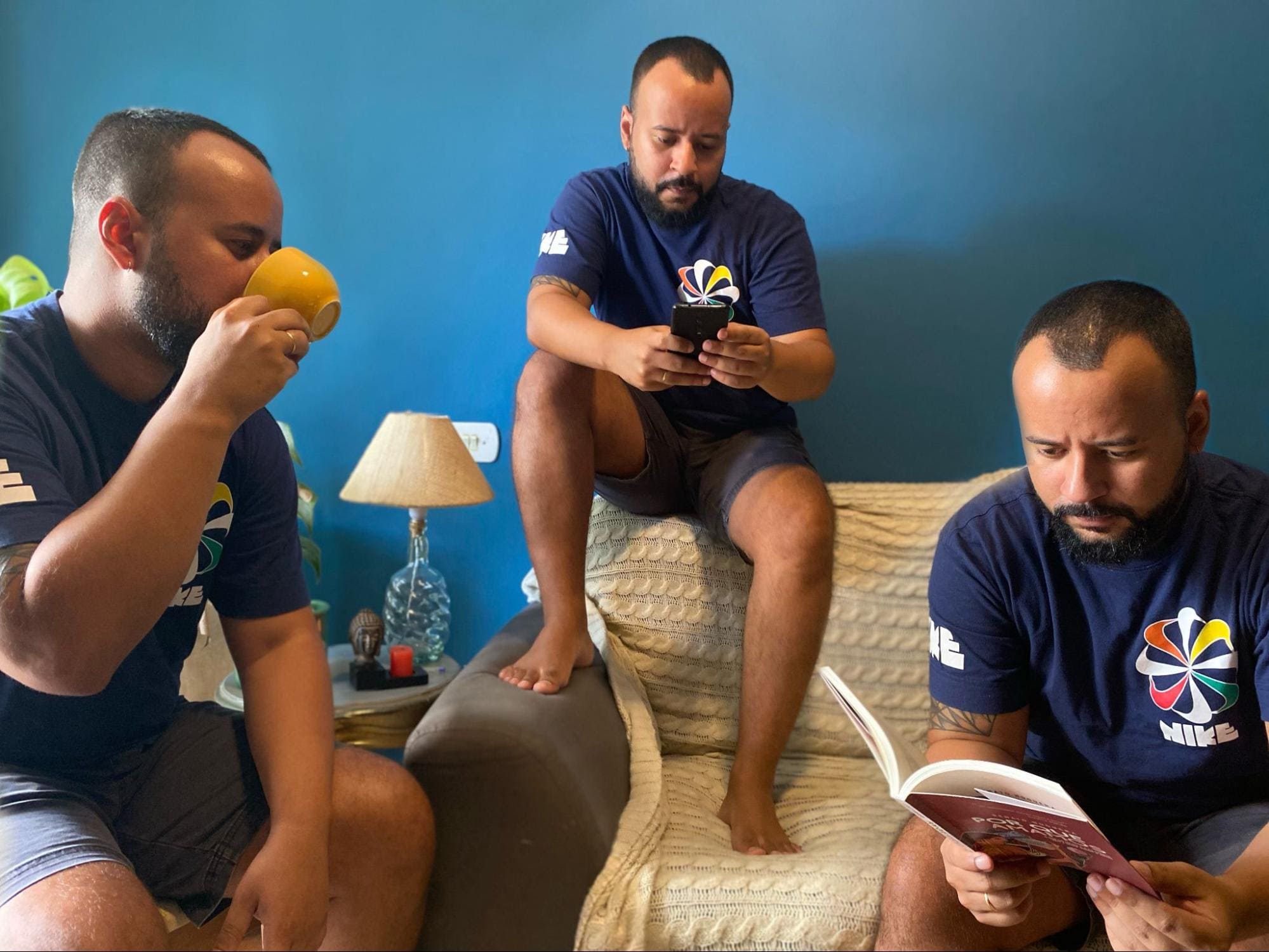 Imagem de uma sala com uma parede azul e um sofá com o personagem aparecendo em três posições diferentes: 1 - homem sentado em uma cadeira do lado esquerdo tomando um café; 2- homem sentado em cima do sofá no meio vendo celular; 3- homem sentado no sofá do lado esquerdo lendo um livro.