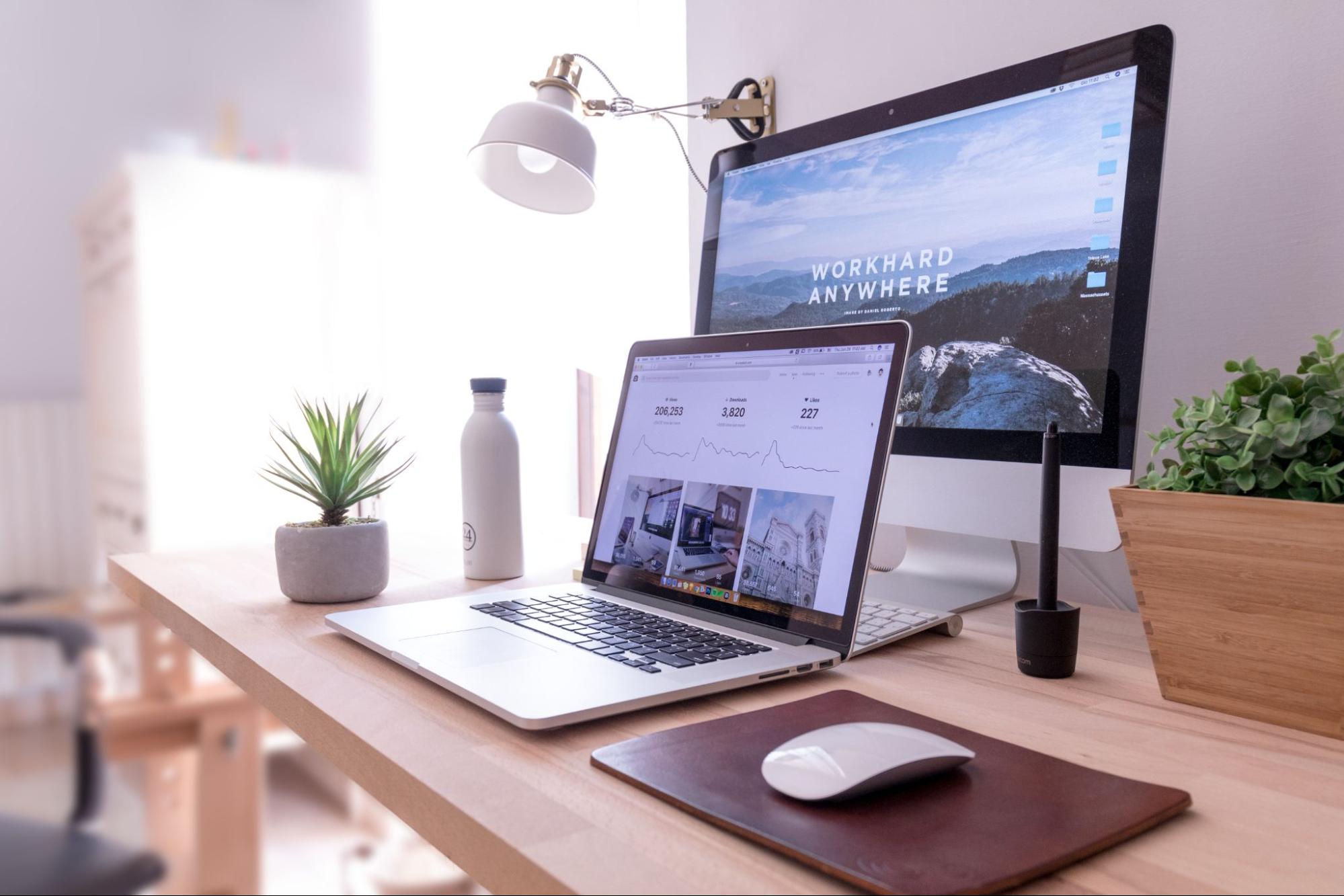 Imagem de uma mesa com uma tela grande, um notebook, um mouse sobre um mousepad, duas plantas pequenas, uma garrafa, um teclado e uma luminária pendurada na parede.