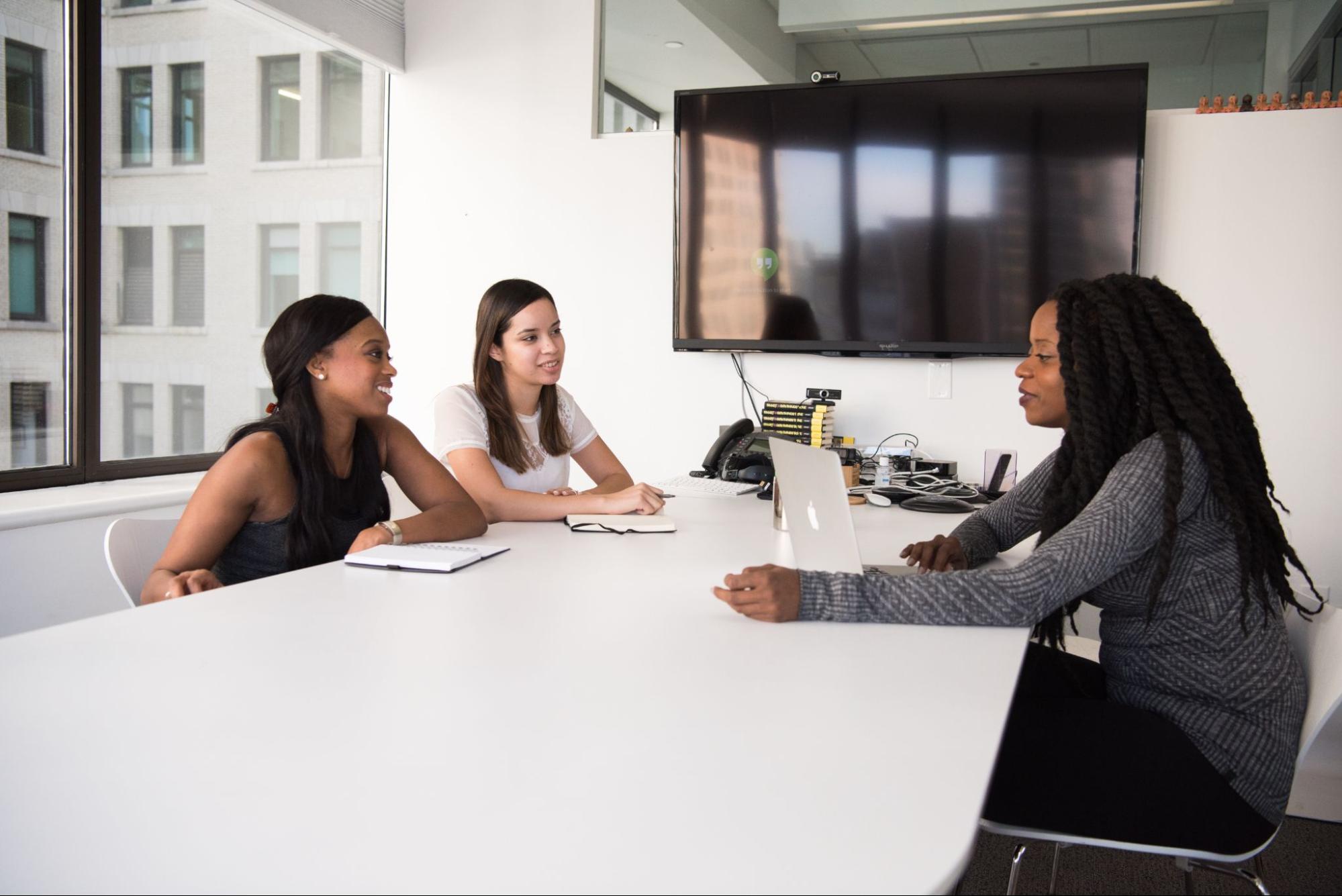 Imagem mostrando três mulheres sentadas à mesa, duas com cadernos de anotação e uma com um notebook. Elas estão em uma sala de paredes brancas na qual há janelas de um lado e uma televisão pendurada do outro.