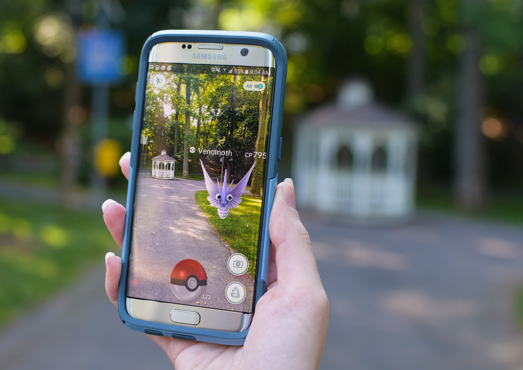 Imagem que mostra uma mão segurando um celular, em um local aberto. Na tela do celular, aparece o jogo Pokemon GO, em realidade aumentada, mostrando além da imagem real, elementos do jogo.