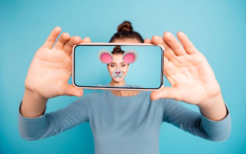 Imagem de uma mulher com um celular sobre o rosto. Nele, há um filtro de snapchat, que mostra a mulher utilizando em sua fotografia um filtro com orelhas e boca de um  rato.