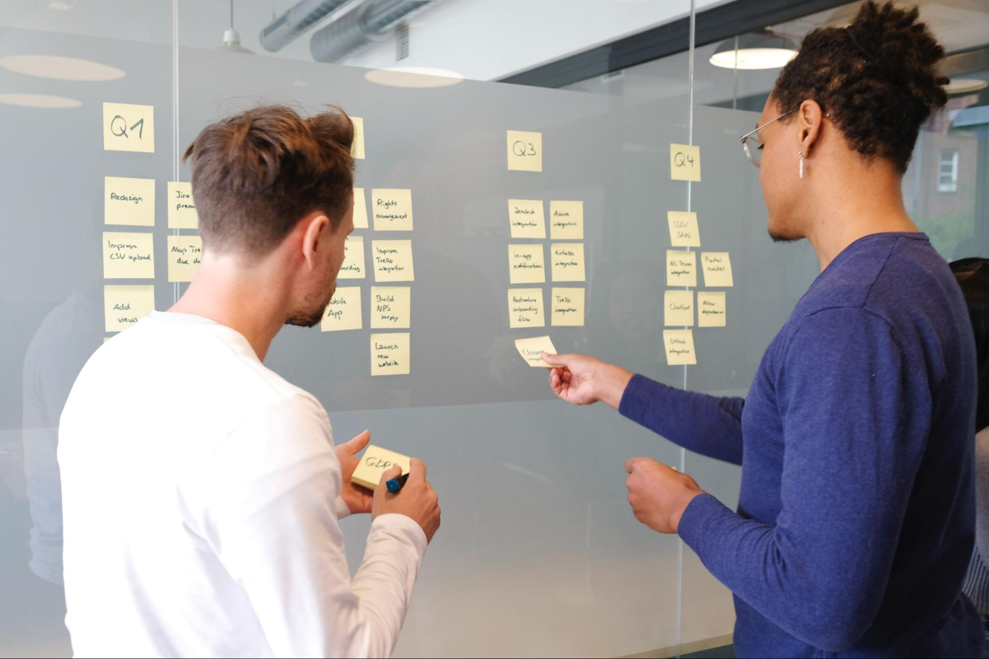 Imagem com dois homens de costas colando post it com diferentes coisas escritas em uma parede de vidro. O homem da esquerda é branco e está usando uma camisa branca e o da direita é negro e está usando uma camisa azul.