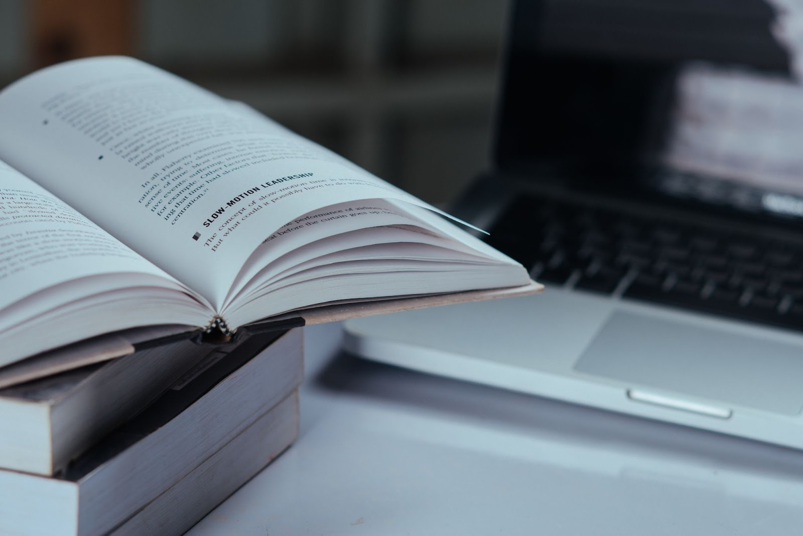 4 livros empilhados, o de cima está aberto mostrando algumas páginas. Atrás dos livros, há um notebook aberto, aparecendo o touchpad, o teclado e a tela, a imagem do notebook está bem desfocada.