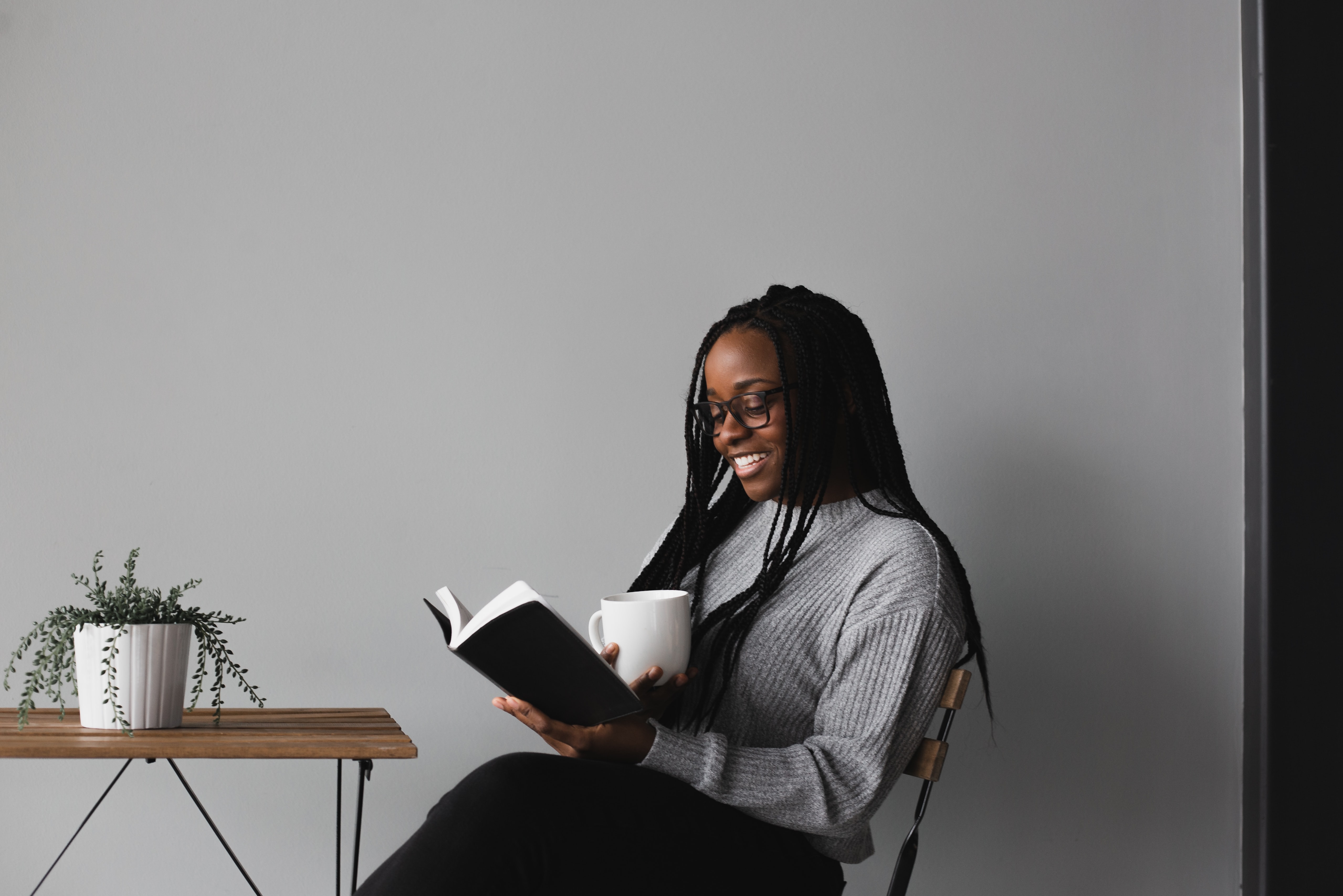 Imagem de uma mulher sentada em uma cadeira e de frente para uma mesa. Na mão direita ela segura um livro e na mão esquerda uma xícara.