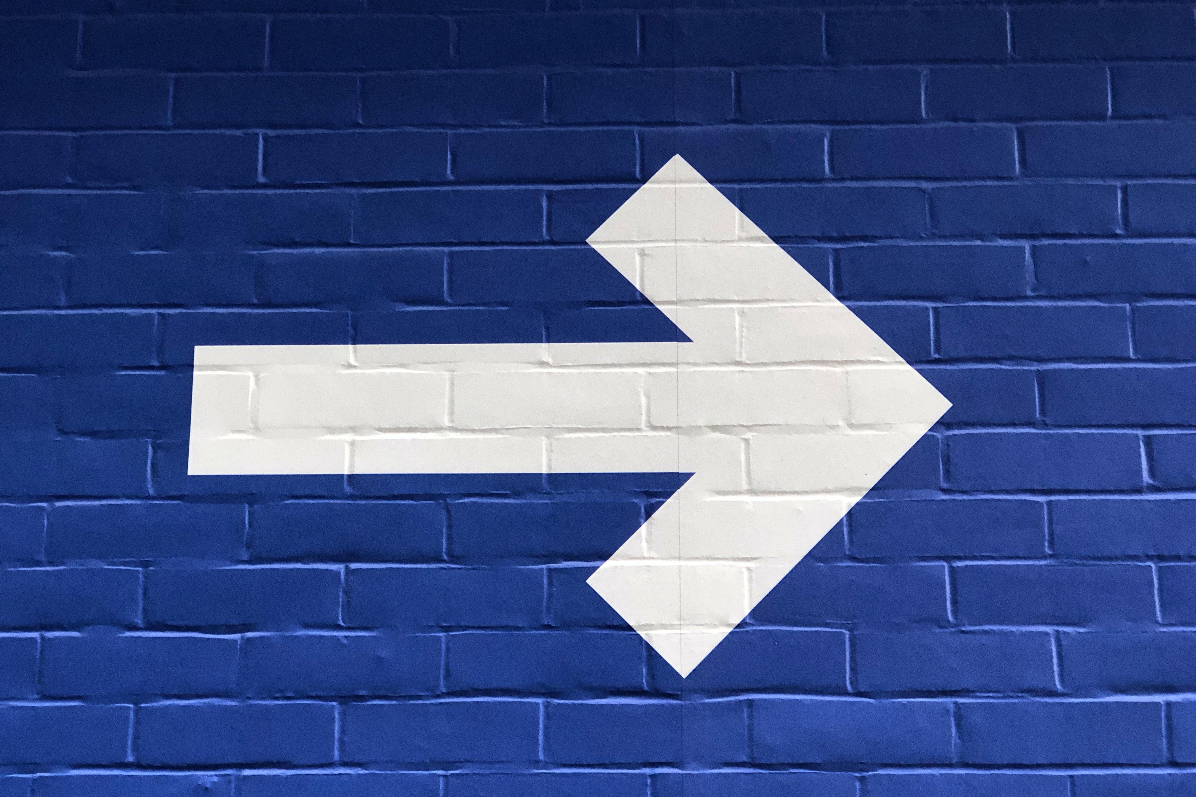 Imagem de uma parede de tijolos pintada de azul e com uma grande seta na cor branca apontada para o lado direito.