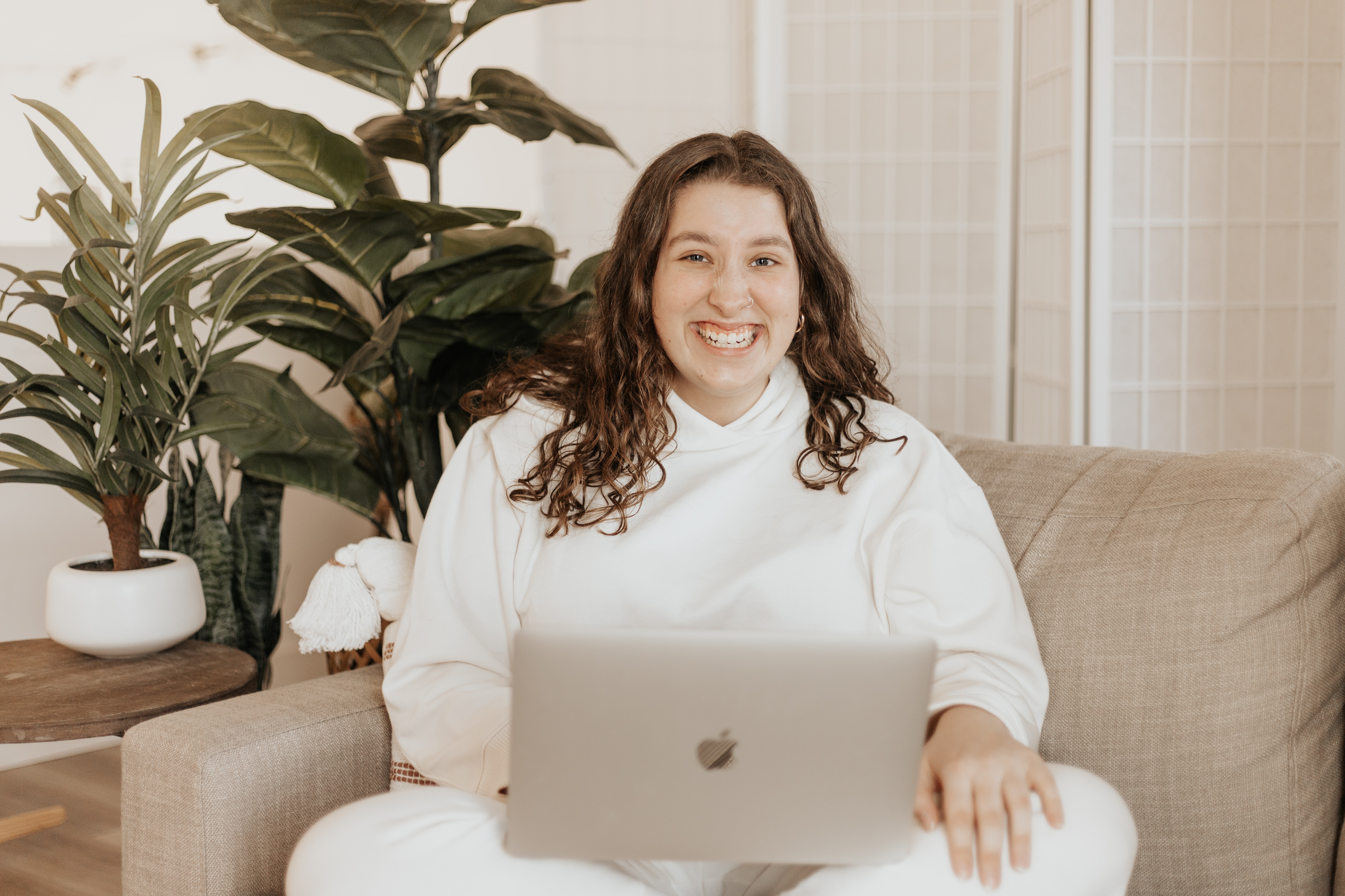 Imagem de uma mulher sentada sobre um sofá e apoiando um notebook sobre as pernas. Ao fundo há algumas plantas, uma mesa pequena e uma divisória para dividir o local em que a mulher está de um outro cômodo.