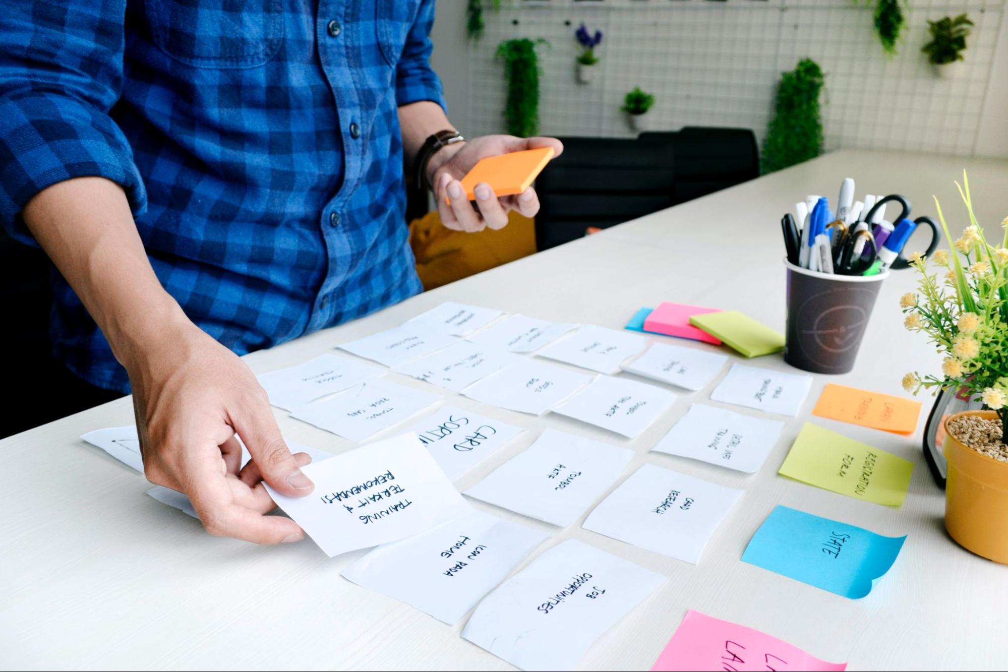 Imagem de uma pessoa organizando cartões de papel e alguns post-its coloridos em cima de uma mesa.