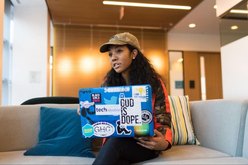 Imagem de uma mulher sentada de pernas cruzadas em um sofá com notebook azul com adesivos colados referentes a tecnologia. A mulher está conversando com outras pessoas que não aparecem na foto.