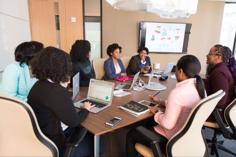 Imagem de uma sala de reuniões com sete mulheres sentadas à mesa. Em cima da mesa tem notebooks, celulares e agendas. Ao fundo da sala tem uma televisão na parede com a apresentação da reunião.