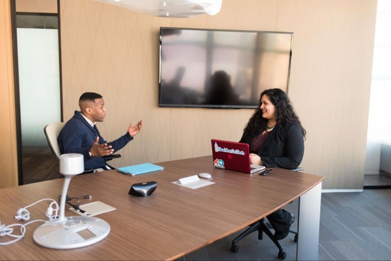 Foto de uma sala de reuniões com um homem e uma mulher sentados à mesa conversando. Na mesa em frente a mulher tem um notebook vermelho aberto, e em frente ao homem uma tablet com capa azul. Na mesa há dispositivos multimídia para reunião, e ao fundo da sala uma televisão desligada.