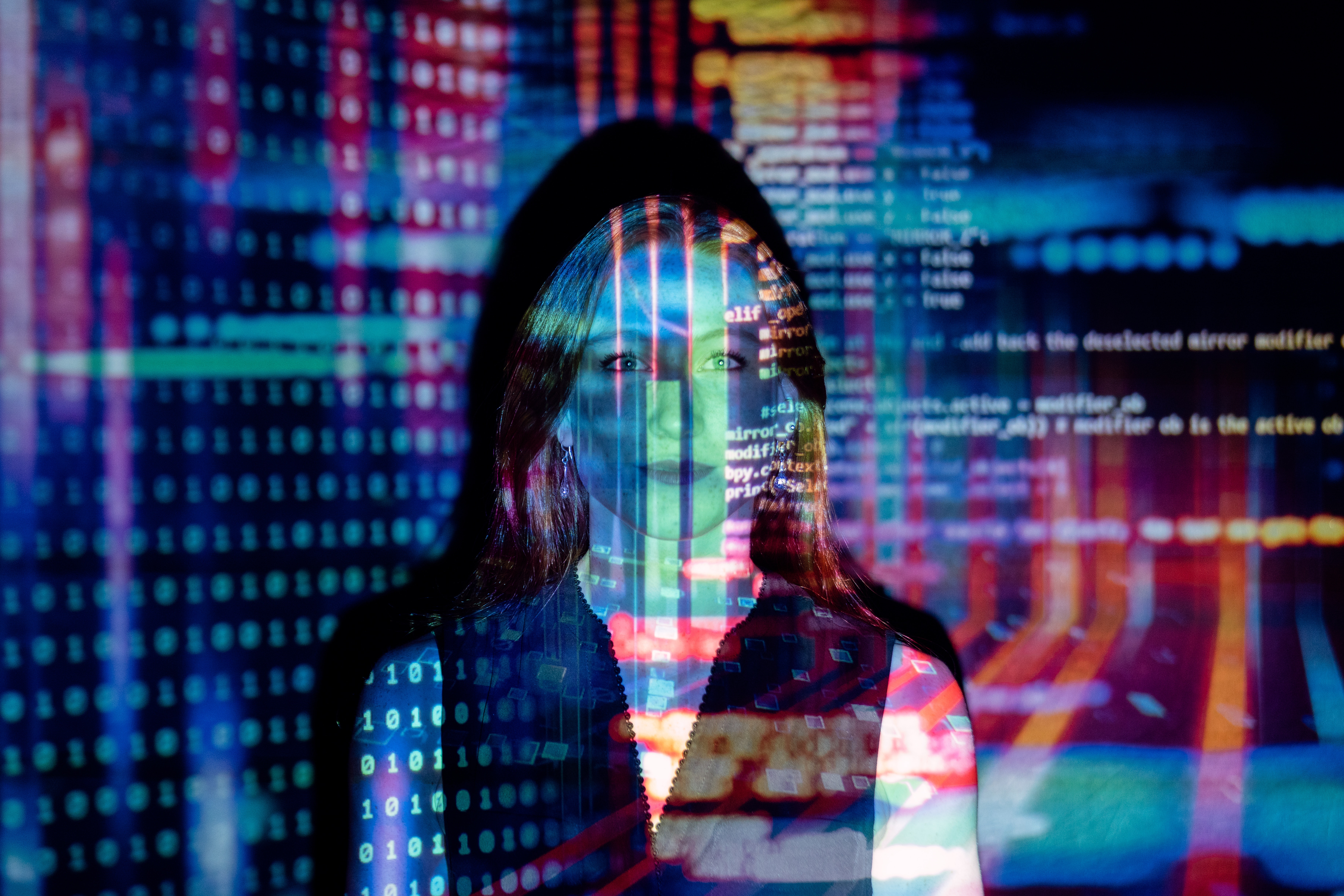 Descrição da imagem: Uma mulher está diante de uma tela de projeção e nela está sendo projetado um emaranhado de números binários e códigos.