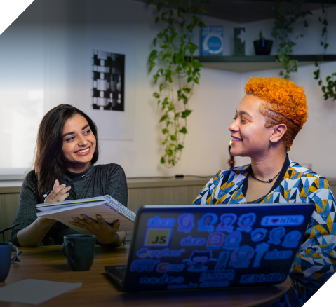 Duas mulheres em frente ao computador conversando enquanto uma mostra algo no caderno para outra