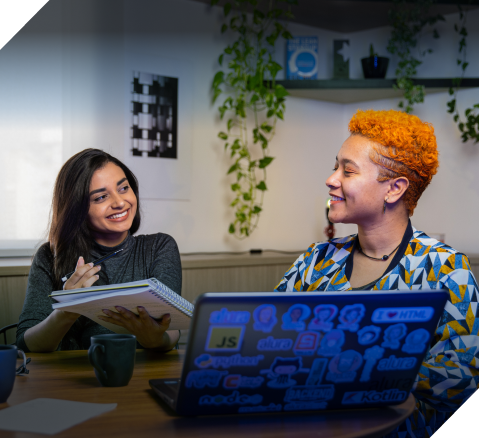 Duas mulheres em frente ao computador conversando enquanto uma mostra algo no caderno para outra