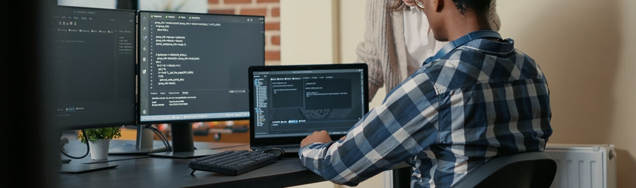 Duas pessoas estão sentadas em um escritório corporativo, trabalhando em suas mesas repletas de várias telas exibindo linhas de código em execução.