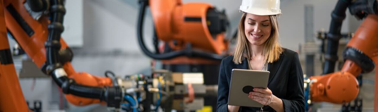 mulher em uma fábrica, próximo a máquinas, mexendo em um tablet, representando a automação industrial