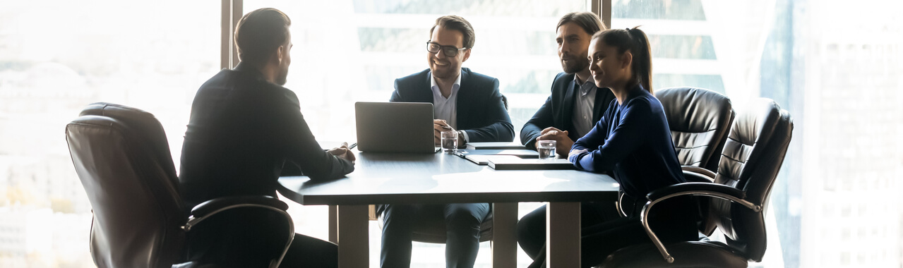 Pessoas diversas profissionais de RH sentadas em uma mesa corporativa com um notebook sobre ela, conversando e discutindo descontraidamente. 