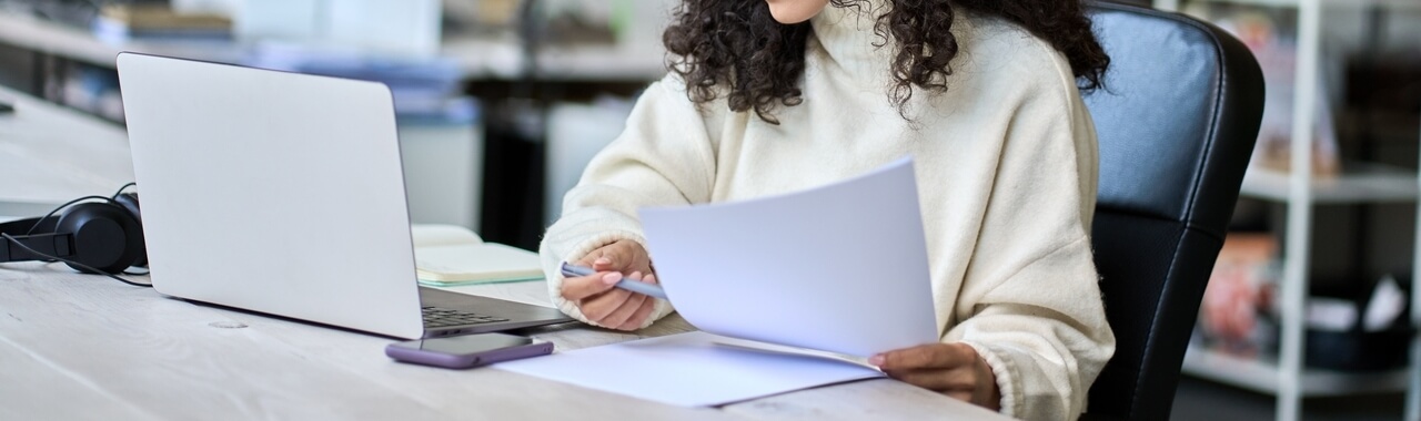 A imagem retrata uma jovem mulher latina profissional de RH sentada em uma mesa de escritório corporativo. Ela segura uma pequena pilha de papéis na mão enquanto verifica algumas informações em seu notebook. Essa imagem representa como o ChatGPT no RH pode ser usado para apoiar e facilitar as tarefas diárias nesse ambiente de trabalho.