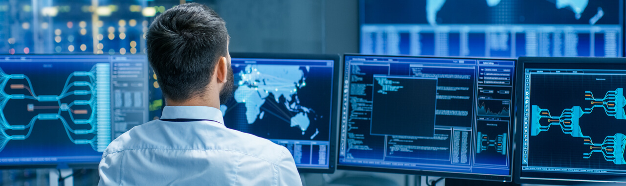 Uma pessoa sentada em frente à três telas de computador, vestindo roupa social,  e observando as informações nas telas. As telas mostram gráficos e dados representando a prática de cibersegurança.
