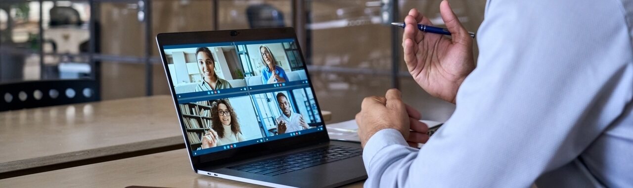 Na imagem, podemos ver uma pessoa líder de tecnologia sentada em uma mesa em um escritório corporativo moderno, focado em uma videochamada com sua equipe. Essa imagem representa o ato de desenvolver pessoas em tecnologia, através da liderança e do compartilhamento de conhecimentos e experiências.