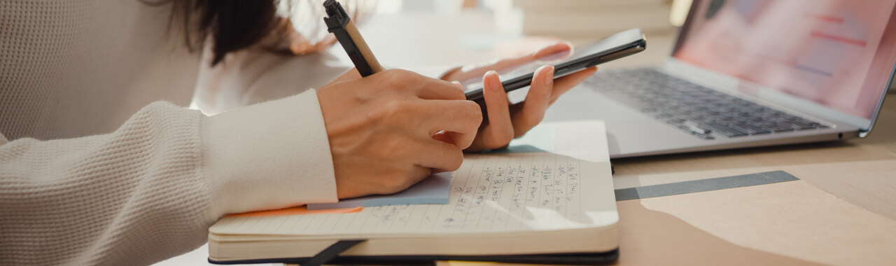 Pessoa com cabelos longos e escuros criando um cronograma de estudo em uma agenda, enquanto usa o celular ao mesmo tempo.