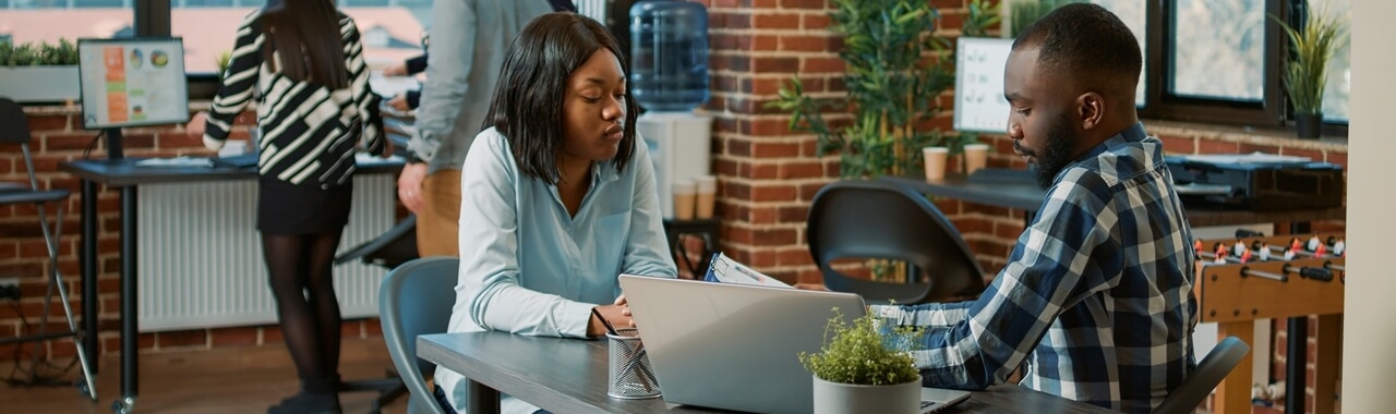 Homem e mulher conversandoem uma mesa corporativa com um notebook e algumas papeladas.