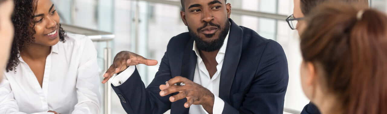 um homem negro de terno falando em uma mesa com outros executivos, representando a ideia de treinamento e cursos para lideranças