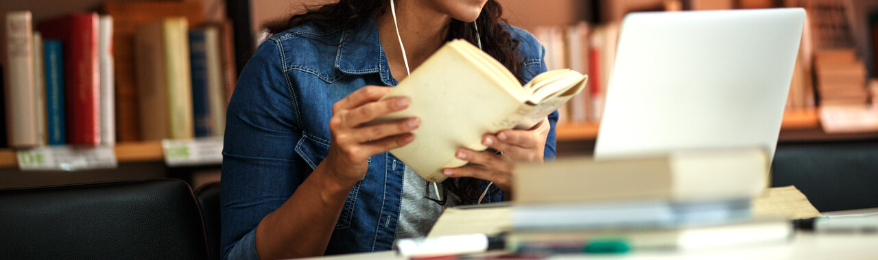 Na imagem, pode-se ver uma pessoa estudante sentada, imersa em um livro que segura com as duas mãos, enquanto assiste algo em um notebook aberto em sua frente. Com alguns livros empilhados na sua mesa, também é possível notar uma estante de livros no fundo, o que reforça a ideia de que ela está em uma biblioteca estudando, simbolizando a ideia de engajamento no aprendizado.