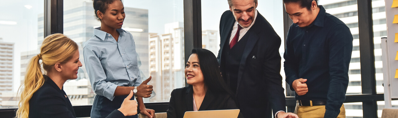 Na imagem de um escritório corporativo, vemos cinco pessoas em uma mesa. Duas mulheres estão interagindo animadamente, uma sentada e outra em pé, trocando sinais de aprovação para a terceira mulher que também está sentada. Ao lado delas, dois homens estão de pé trocando entre si. A imagem representa o ato de se ter feedbacks 360 com a equipe